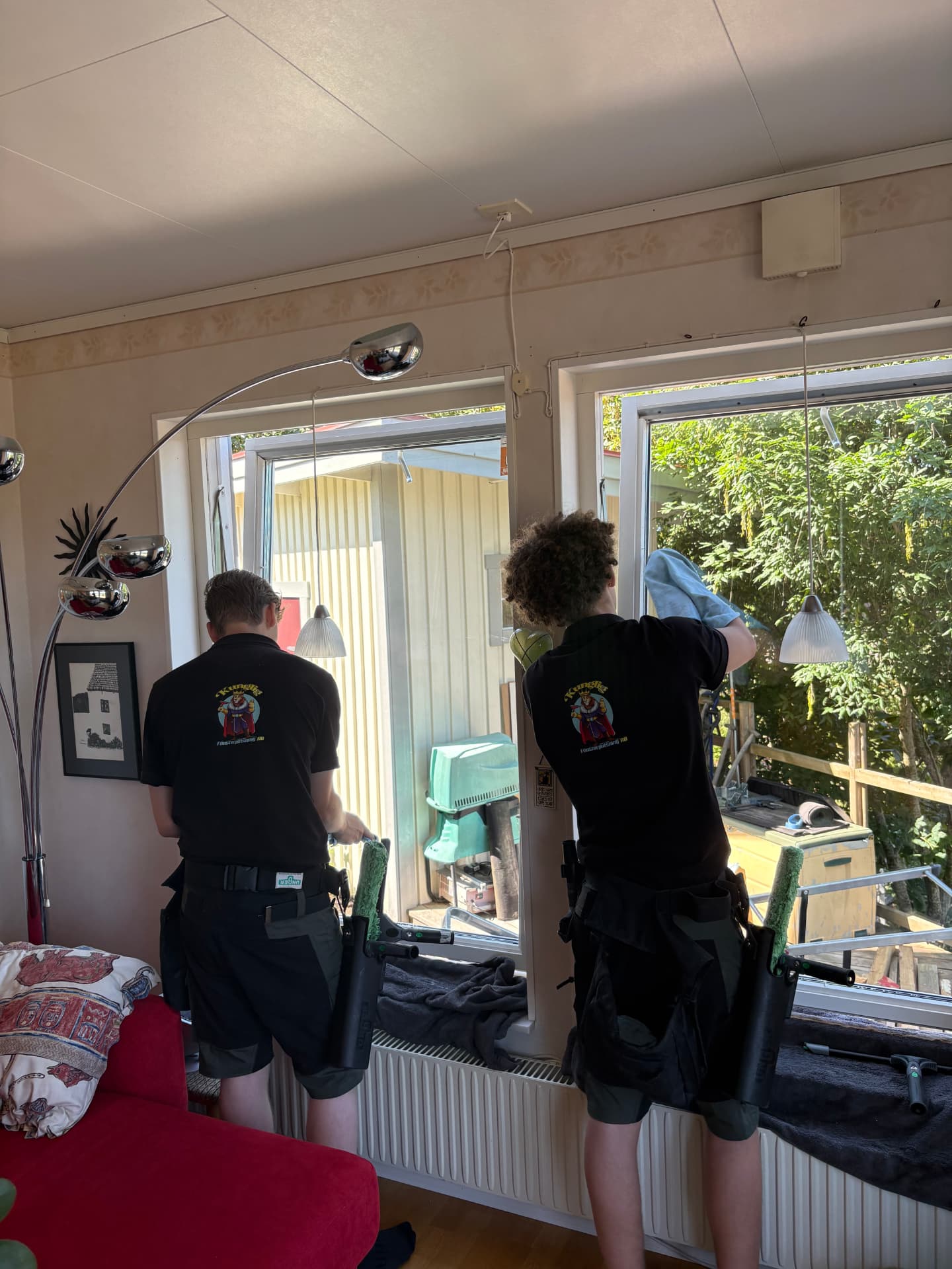 Två fönsterputsare i svart arbetskläder med företagets märke putsar fönster inifrån en villa